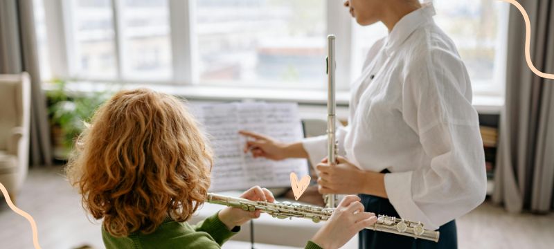 Red-haired child plays the flute while a teacher in a white shirt points at the music stand